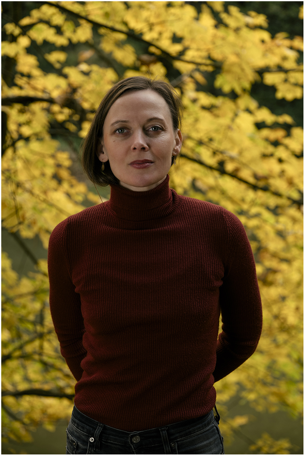 Goldfuchs stands at the edge of a lake in nature, wearing a red sweater with arms crossed behind her back, in front of yellow autumn leaves.