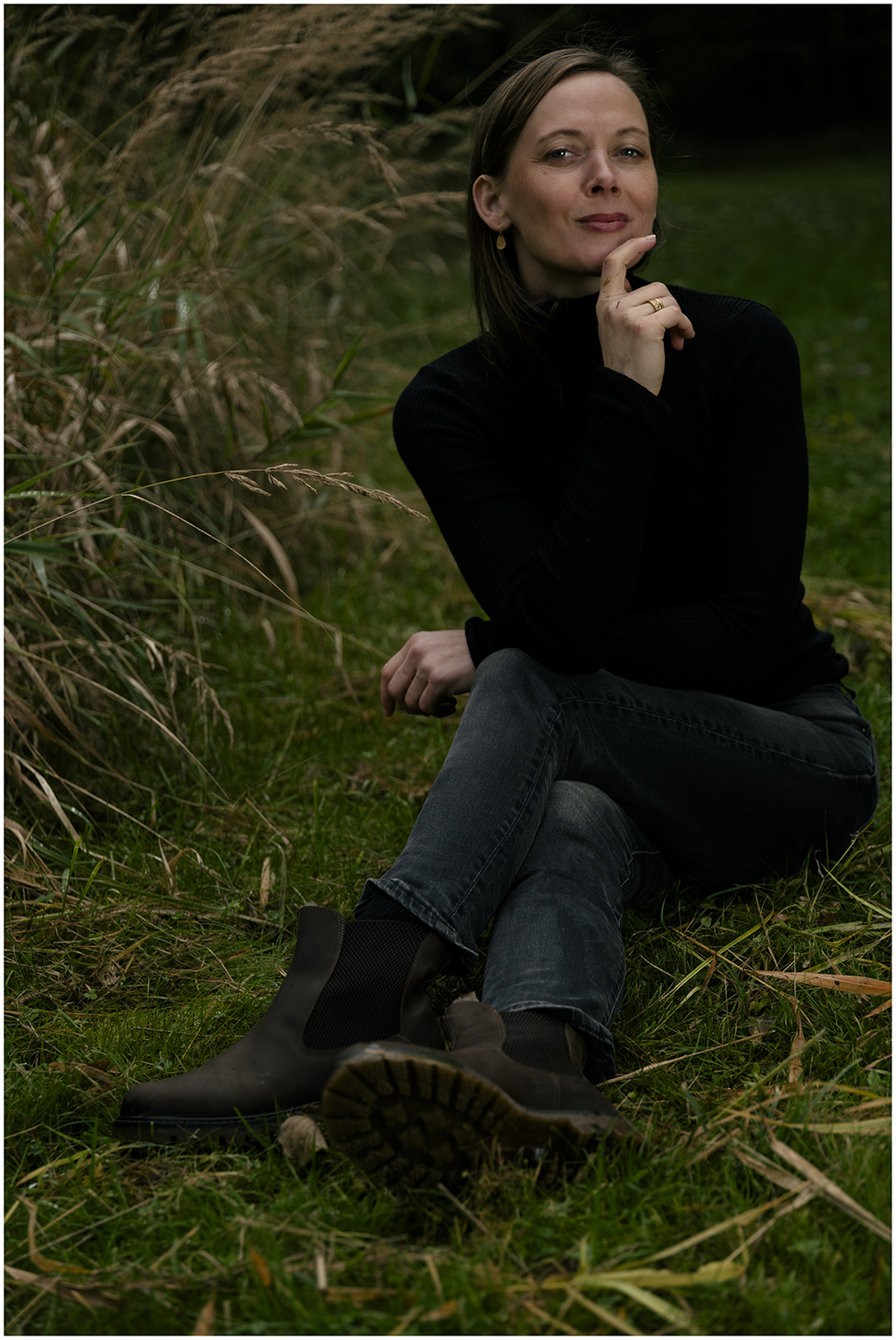 Goldfuchs sits in a meadow in nature, resting her right arm with her index finger on her chin, wearing a black top and smiling.