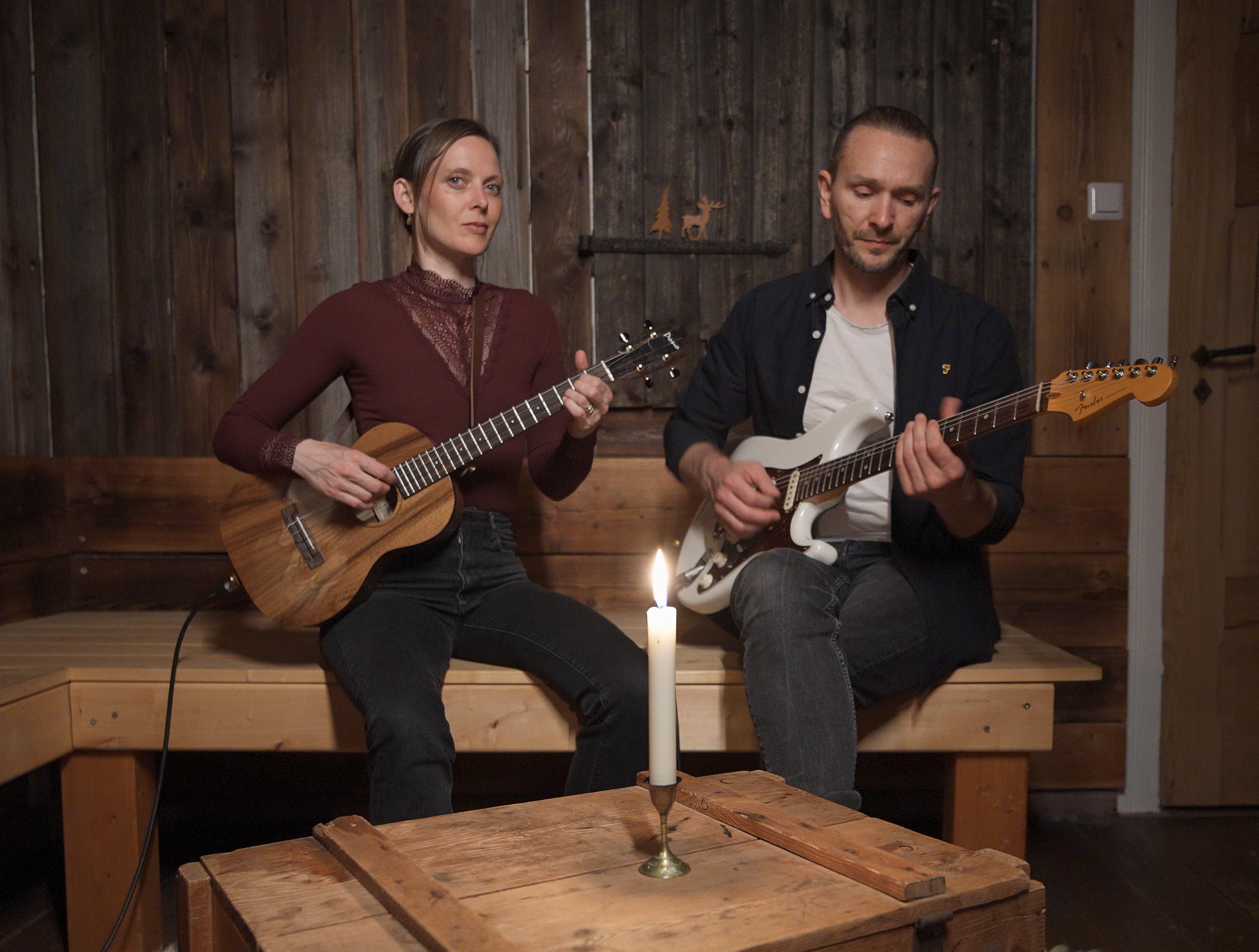 Goldfuchs and Leon Krasmann sit on a rustic bench, each holding their instruments, with a candle glowing in front of them. Goldfuchs wears a burgundy top with black jeans, while Leon Krasmann wears a black shirt over a white undershirt and plays his Fender guitar.
